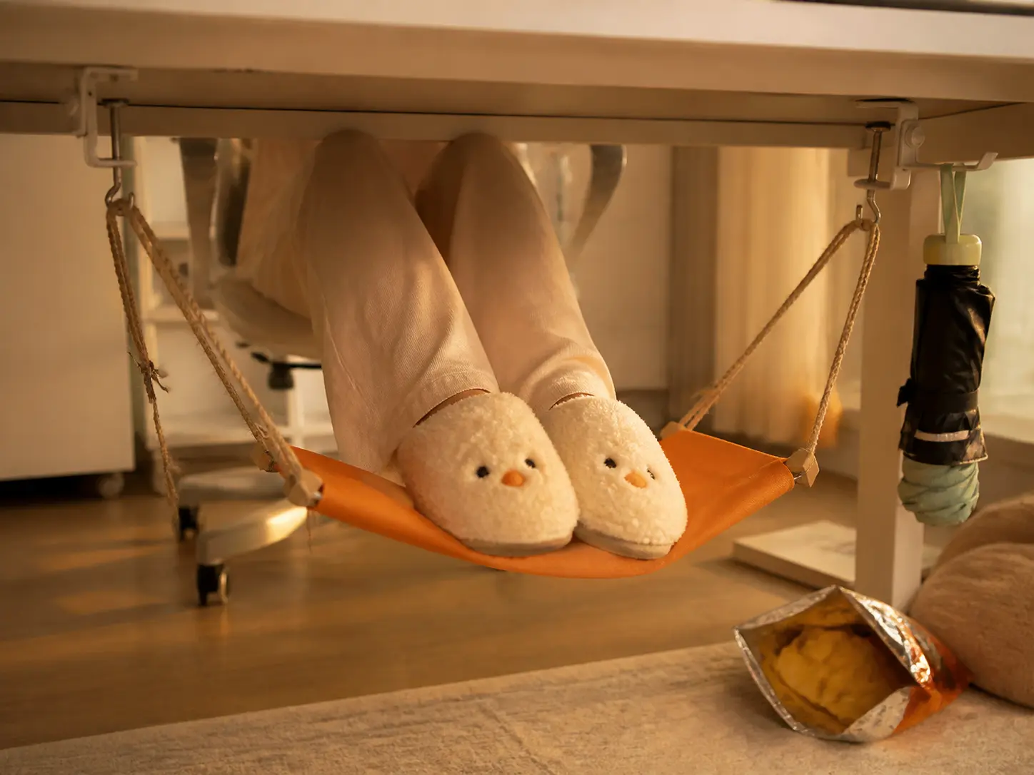 Orange canvas foot hammock hanging beneath a white desk with slippers resting inside